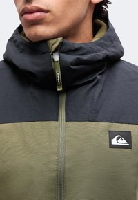 Gros plan d'une personne portant une veste à capuche verte et noire avec une fermeture éclair sur le devant et un écusson avec un logo vague sur la poitrine.