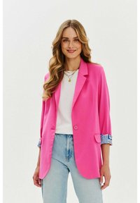 Ružový blazer s prispôsobeným strihom, jedným gombíkom a srolovanými rukávmi, ktorý má modrú vzorovanú látku. Nosený cez biely top a svetlé džínsy.