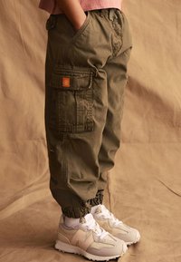 Olivgrön cargobyxor med elastiska cuffar, flera fickor, en patchlogotyp, kombinerade med ljusbeige sneakers och vita strumpor.