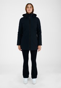 Giacca nera impermeabile con cappuccio, dotata di una zip anteriore e due tasche laterali, abbinata a pantaloni neri e sneakers bianche.