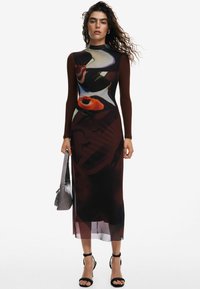 Rochie cu mânecă lungă, de culoare maro închis, dintr-un material imprimat cu modele abstracte. Detaliu transparent la tiv. Modelul ține în mână o geantă gri și poartă sandale negre cu barete.