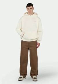 Cremefarbener Hoodie mit Fronttasche, kombiniert mit braunen weiten Hosen; getragen mit weißen und dunklen Schuhen; minimalistisches Design.