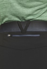Gros plan de la taille d’un pantalon noir avec ceinture élastique à motif et poche zippée, porté avec une chemise verte.