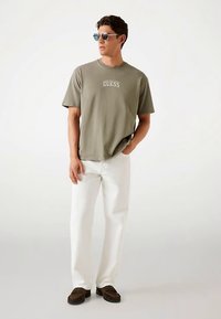 Oliwkowa zielona koszulka z logo "GUESS", zestawiona z białymi spodniami i brązowymi butami wsuwanymi, modelka nosi okulary przeciwsłoneczne, stoi na neutralnym tle.