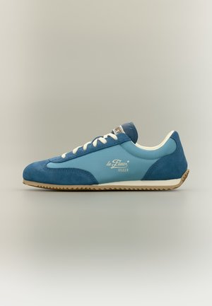 Μπλε και ανοιχτό μπλε sneaker χαμηλού τύπου με λευκά κορδόνια, μπεζ σόλα και σήμανση "le Fleur Jogger" στην πλευρά.