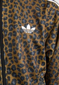 Hnedý kabát s leopardím vzorom a čiernymi akcentmi, s bielym logom a detailom zipsu. Vyrobený z hladkej, mierne textúrovanej látky.