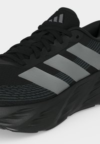 Μαύρο αθλητικό παπούτσι με γκρι ρίγες Adidas, μαύρα κορδόνια, υφή στη σόλα και επενδεδυμένο γιακά ορατά σε λευκό φόντο.