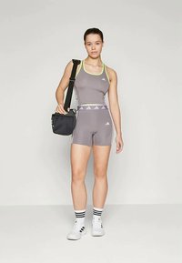 Pelēks sporta tvertnes tops ar neona dzelteniem akcentiem, pārī ar atbilstošām šortiem. Figūra tur melnu pleca somu, valkā baltas kurpes un svītrainas zeķes.