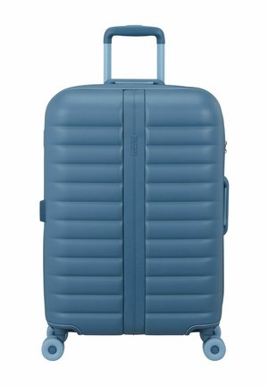Valise rigide bleue avec des rainures horizontales, poignée rétractable, quatre roulettes pivotantes et poignée latérale.
