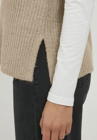 Beige värillinen ribbineule, jossa on sivuhalkio, yhdistetty valkoiseen pitkähihainen paitaan. Näkyvissä tumman denimin päällä korostaen tekstuuria ja kerroksellisuutta.