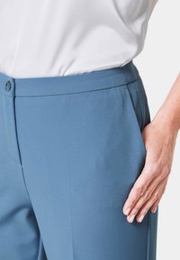 Maßgeschneiderte blaue Hose aus glattem Stoff, mit flachem Bund, Taschen und einem schlichten Taillenbund. Hand ruht an der Seite der Hose.