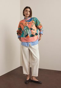 Trui met een levendig bloemenpatroon in roze, blauw en groen, gecombineerd met een lichtblauw overhemd met kraag. Gecombineerd met crèmekleurige wijde broekspijpen en zwarte platte schoenen.