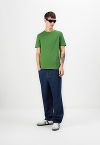 Young man wearing black sunglasses, green short-sleeve shirt, loose dark blue jeans, white sneakers, standing against plain white background.