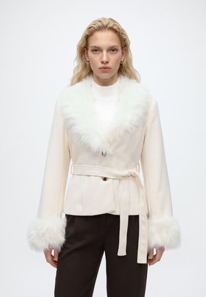 Femme blonde portant un manteau blanc ceinturé avec un col et des poignets blancs duveteux, associée à un pantalon foncé, debout devant un fond uni.