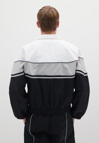 Kleurgeblokte jas met een witte bovenkant, grijze middenpartij en zwarte onderkant. Elastische tailleband en manchetten. Lichtgewicht stoftextuur.