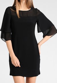 Mustassa lyhyessä mekossa on läpinäkyvät, kellomaiset hihat ja pyöreä pääntie. Kangas on sileä, kiinteä tekstuuri ja yksinkertainen muotoilu.