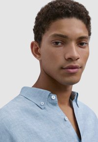 Light blue, button-up shirt with a textured fabric, featuring a modern collar and two buttons at the neckline. Close-up view of the shirt model.
