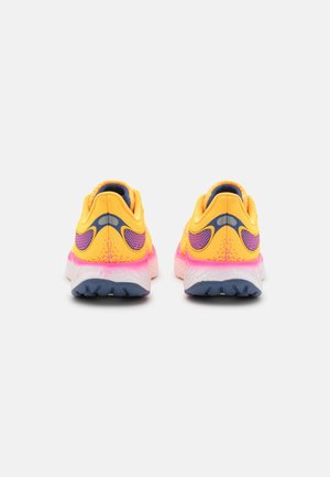 Par de zapatos deportivos amarillos con detalles en rosa y suelas azul oscuro, mostrados desde atrás sobre un fondo claro y liso.