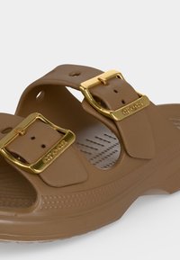 Καφέ σανδάλι με δύο ρυθμιζόμενες χρυσές αγκράφες χαραγμένες με "Crocs" και υφασμένο πέλμα για άνεση και κράτημα.