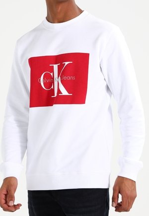 Mężczyzna w białej bluzie z czerwoną kwadratową grafiką i białym logo "Calvin Klein Jeans", w czarnych spodniach, stojący na jednolitym tle.