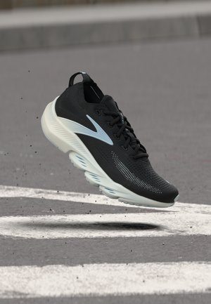 Černo-bílá atletická bota se světle modrými detaily, ve vzduchu nad asfaltem s bílými dopravními značkami a malými rozptýlenými nečistotami.