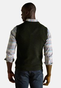 Muž otočený chrbtom, oblečený v tmavozelenom bezrukávovom svetri cez farebnú károvanú košeľu s vyhrnutými rukávmi a tmavými džínami.