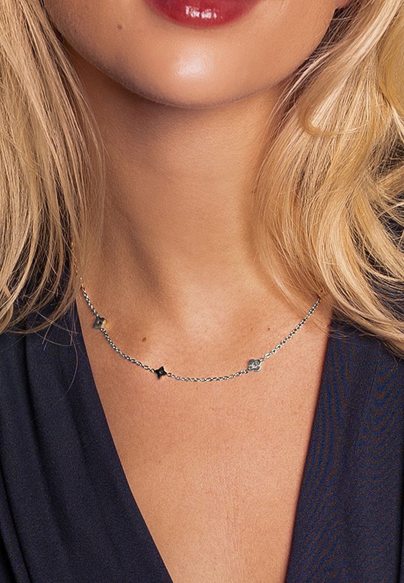 Collier en chaîne argentée avec de petits accents géométriques de formes et finitions variées, présenté sur un fond en tissu foncé.