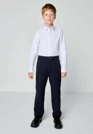 Junger Junge steht und trägt ein weißes Hemd mit Knöpfen, dunkle Hosen und schwarze Schuhe vor einem schlichten grauen Hintergrund.