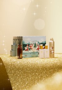 Geschenkset für die Feiertage mit einer dekorativen Box, die ein verschneites Dorf zeigt, einem braunen Glas-Raumbeduftungsdiffusor und verschiedenen weißen und goldenen Flaschen auf einer glitzernden Oberfläche.