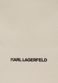 Bombažna kanvas tkanina v svetlem bež odtenku s poudarjenim črnim besedilom "KARL LAGERFELD" na sredini, matirana tekstura z enakomernim tkalskim vzorcem.