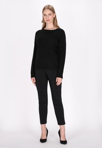 Musta neulepaita, jossa on pyöreä kaula-aukko ja pieni tasku, yhdistettynä slim fit -mustaakin housujen ja mustien korkokenkien kanssa. Sileä pinta, istuva muotoilu.