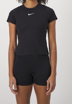 Femme portant une tenue de sport noire ajustée de Nike composée d'un top à manches courtes et d'un short taille haute, sur un fond uni.