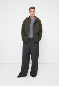 Olivovo zelená bunda s kapucňou, šedé polo tričko pod ňou a voľné tmavošedé nohavice. Hladké textúry, detaily na patentkách a bočné vrecká.