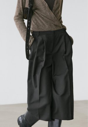 Czarne, wysokowzystające spodnie z zakładkami, wykonane z gładkiej, lekko błyszczącej tkaniny, zestawione z brązowym kardiganem i czarną torebką na pasku.