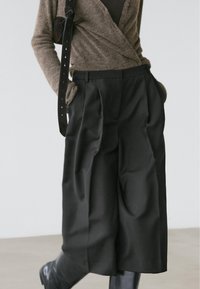 Pantalon noir taille haute, plissé, en tissu lisse et légèrement brillant, associé à un cardigan marron et un sac à bandoulière noir.