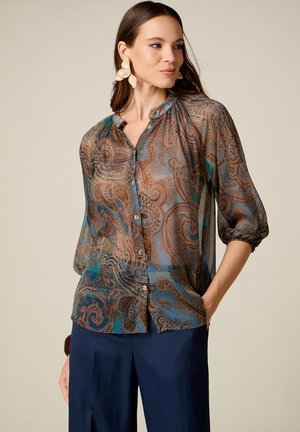 Mujer que lleva una blusa transparente con estampado de cachemira y mangas tres cuartos y pantalones azul oscuro, mirando hacia un lado con las manos en los bolsillos.