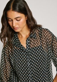Schwarze durchscheinende Bluse mit Knopfleiste, die ein Blumenmuster in Creme, Orange und Blau zeigt, mit einem Kragen und einem lockeren Schnitt.