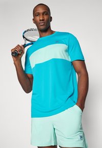 Türkises Sportshirt mit einem hellblauen horizontalen Streifen, kombiniert mit mintgrünen Shorts. Mit Logo auf Shirt und Shorts.