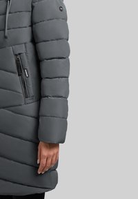 Geaca puf gri cu un design matlasat, având un buzunar cu fermoar și glugă. Materialul este neted și izolat pentru căldură.