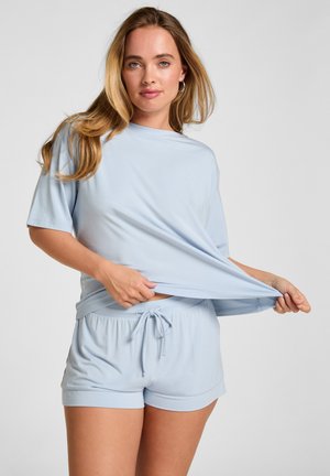 Vrouw met lang blond haar, gekleed in een lichtblauw loszittend T-shirt en korte broek, die het T-shirt iets naar de zijkant trekt.