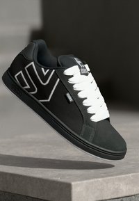 Schwarze und graue Sneaker mit weißen Schnürsenkeln und Etnies-Logo, auf einer Betonoberfläche platziert mit neutral verschwommenem Hintergrund.