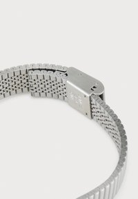 Montre en acier inoxydable avec un bracelet en maille, dotée d'une fermeture à clapet et d'une finition lisse et polie. Gravure visible sur le clapet.