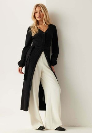 Lyshåret kvinde iført en lang sort cardigan med knapper og bælte over brede hvide bukser og sorte sko, stående op ad en hvid væg.