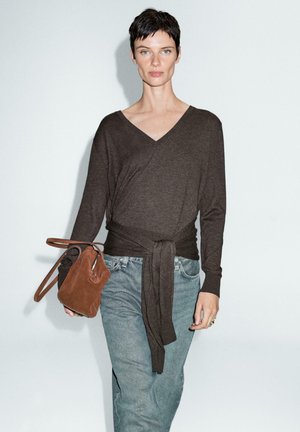 Maglione marrone a maniche lunghe con vita legata. Indossato con jeans blu sbiaditi. Tiene una piccola borsa in suede marrone. Sfondo semplice.