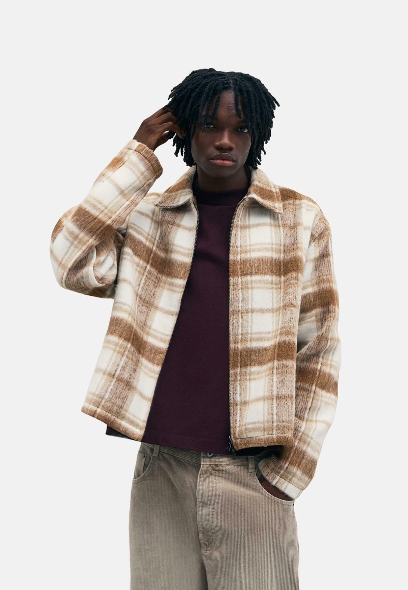 Hnedo-krémová károvaná fleecová bunda s golierom, zipsom na prednej strane a bočnými vreckami, nosená cez tmavú košeľu a béžové kordové nohavice.