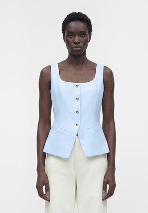 Model feminin purtând o vestă albastru deschis fără mâneci cu nasturi și pantaloni albi cu talie înaltă pe un fundal simplu.