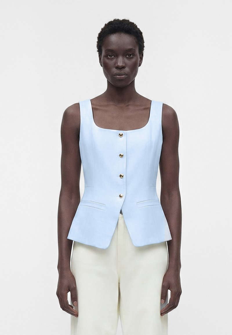 Γυναίκα μοντέλο που φοράει γαλάζιο αμάνικο γιλέκο με κουμπιά και ψηλόμεσο λευκό παντελόνι μπροστά σε απλό φόντο.