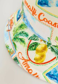 Chapeau seau en tissu à motif avec des citrons, des feuilles de palmier et des scènes côtières. Fond beige clair avec des couleurs vives et des détails de couture.