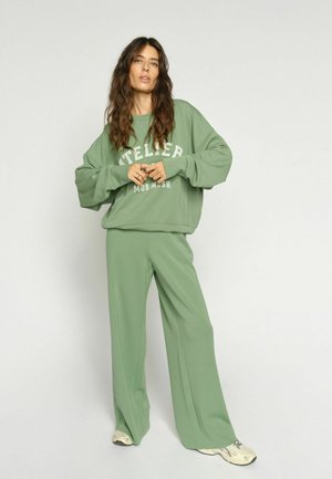 Nő laza, világos zöld pulóverben, amin az "ATELIER MOS MOSH" felirat látható, és hozzá illő, bő szárú nadrágban pózol egy egyszínű háttér előtt.