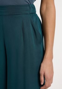 Pantaloni a gamba larga in teal con tessuto liscio, vita elastica e tasche laterali. Presentano delicate pieghe vicino alla vita.